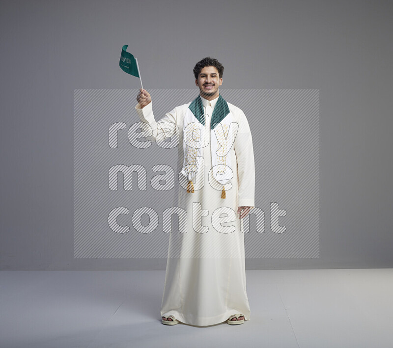 A Saudi man standing wearing thob and saudi flag scarf and holding small saudi flag on gray background