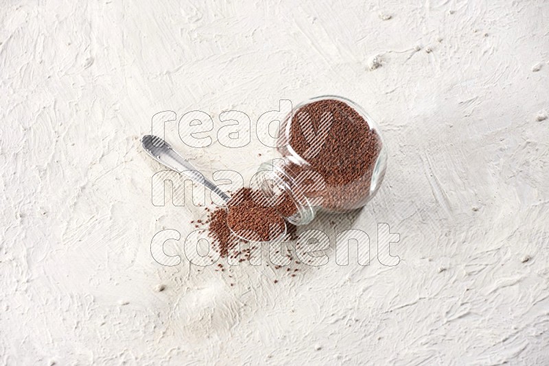 A glass spice jar and a metal spoon full of garden cress seeds and jar is flipped with fallen seeds on a textured white flooring