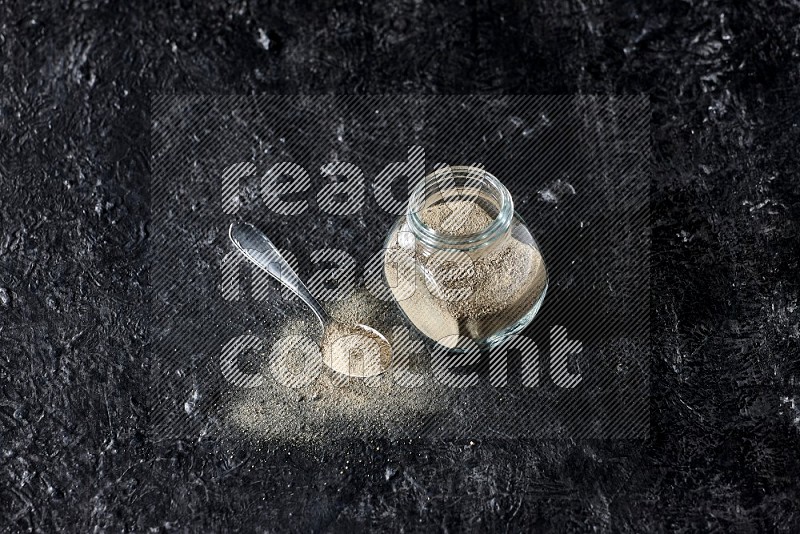 Herbal glass jar and metal spoon full of white pepper powder on textured black flooring