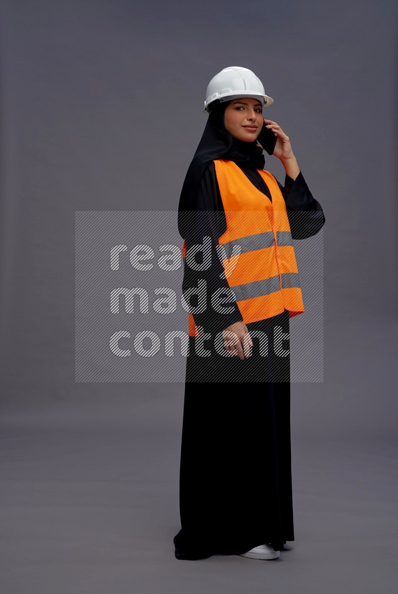 Saudi woman wearing Abaya with engineer vest standing talking on phone on gray background