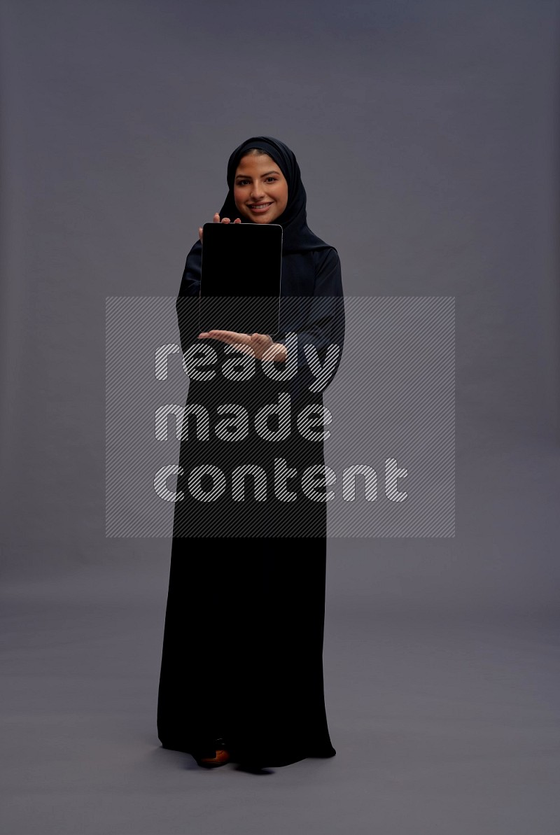 Saudi woman wearing Abaya standing showing tablet to camera on gray background