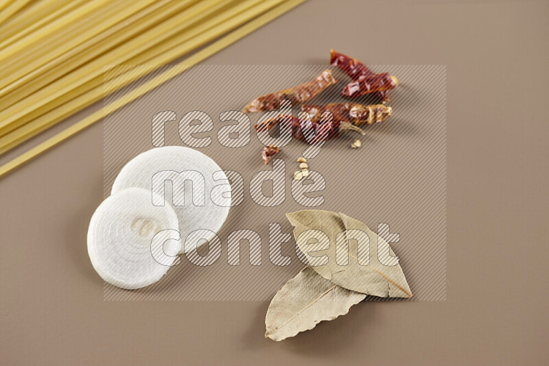 Raw pasta with different ingredients such as cherry tomatoes, garlic, onions, red chilis, black pepper, white pepper, bay laurel leaves, rosemary and cardamom on beige background