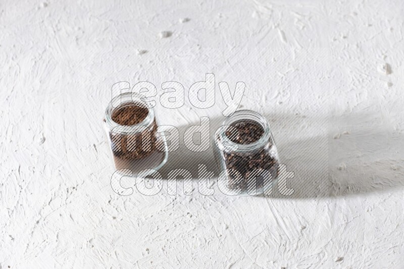 2 glass jars full of cloves powder and cloves grains on a textured white flooring