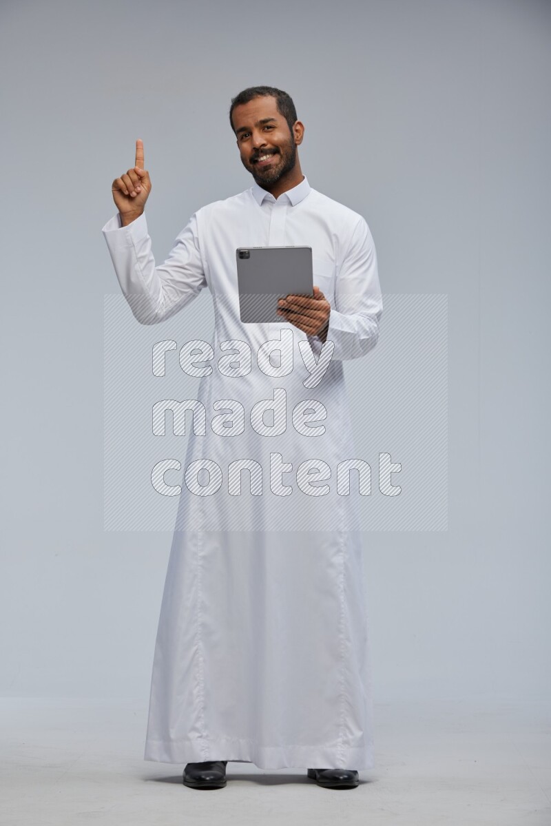 Saudi man wearing Thob standing working on tablet on Gray background