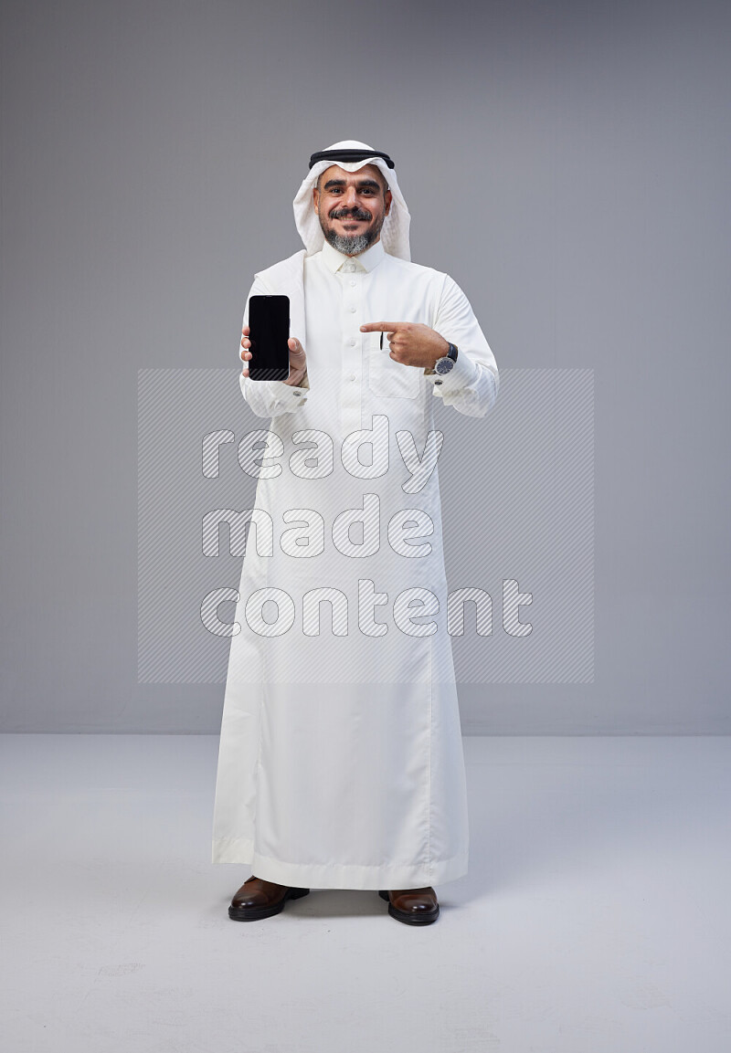 Saudi man Wearing Thob and white Shomag standing showing phone to camera on Gray background