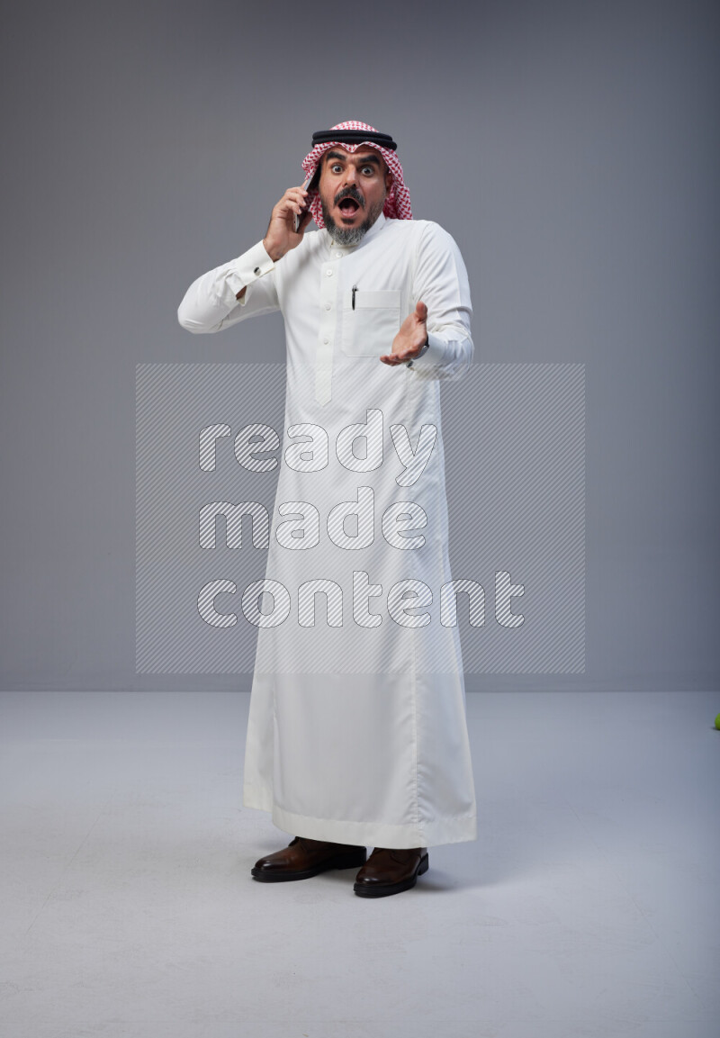 Saudi man Wearing Thob and red Shomag standing talking on phone on Gray background