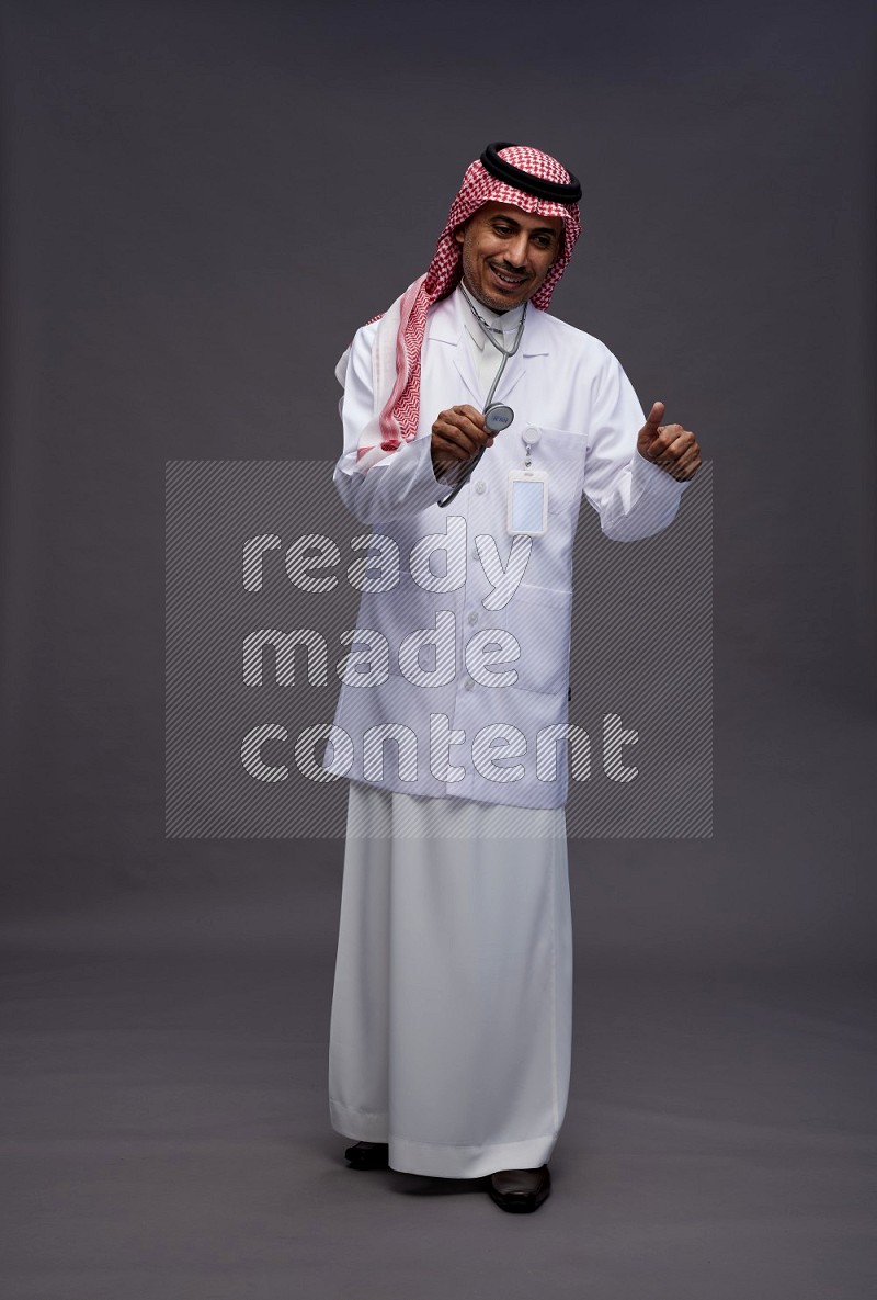 Saudi man wearing thob with lab coat and shomag with pocket employee badge with stethoscope standing interacting with the camera on gray background