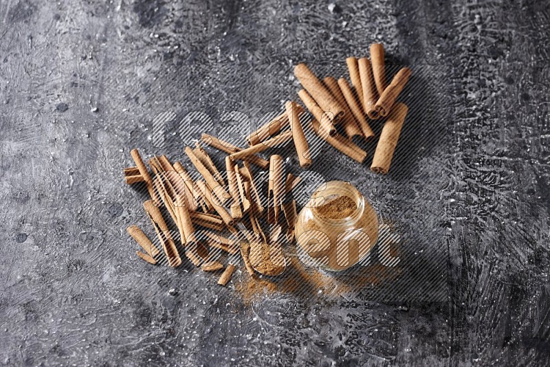 Herbal glass jar and a metal spoon full of cinnamon powder surrounded by cinnamon sticks on textured black background