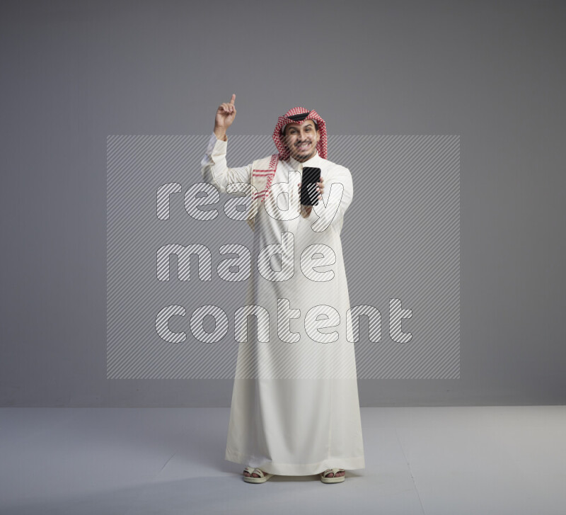 A Saudi man standing wearing thob and red shomag showing phone to camera on gray background