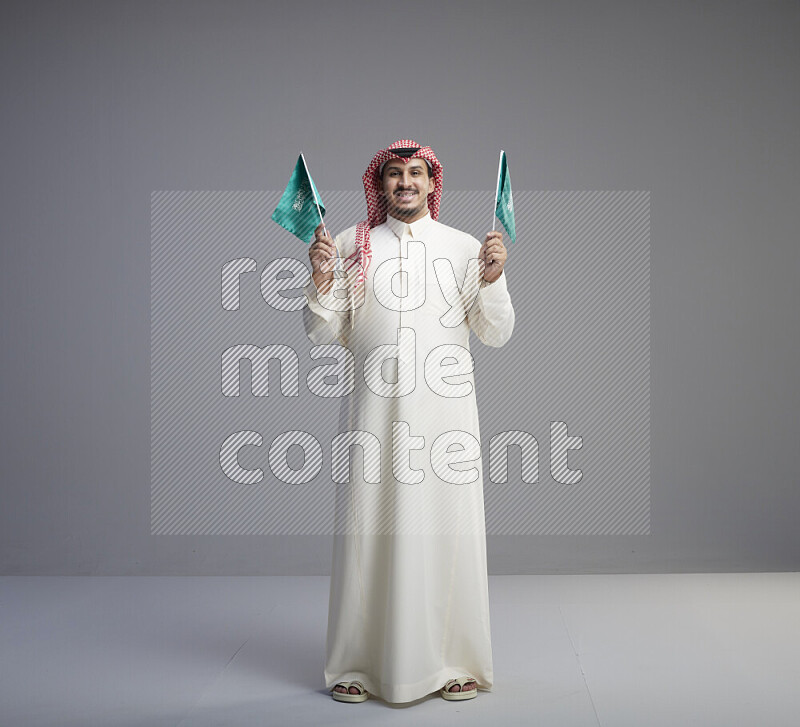 رجل سعودي يرتدي ثوب ابيض وشماغ احمر يمسك بعلمين صغيرين للمملكة العربية السعودية
