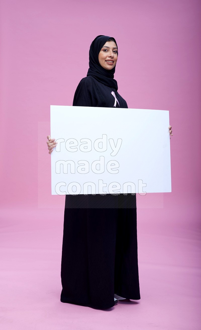 Saudi woman wearing pink ribbon on Abaya standing holding board on pink background