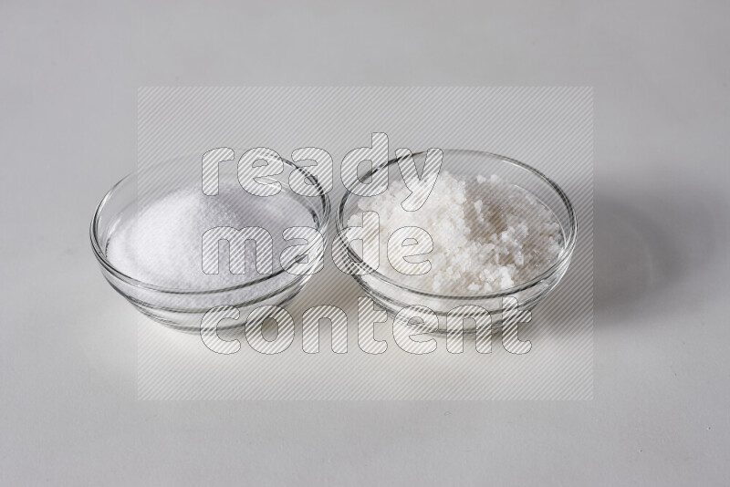 2 glass bowls one is filled with table salt and the other with coarse sea salt on white background