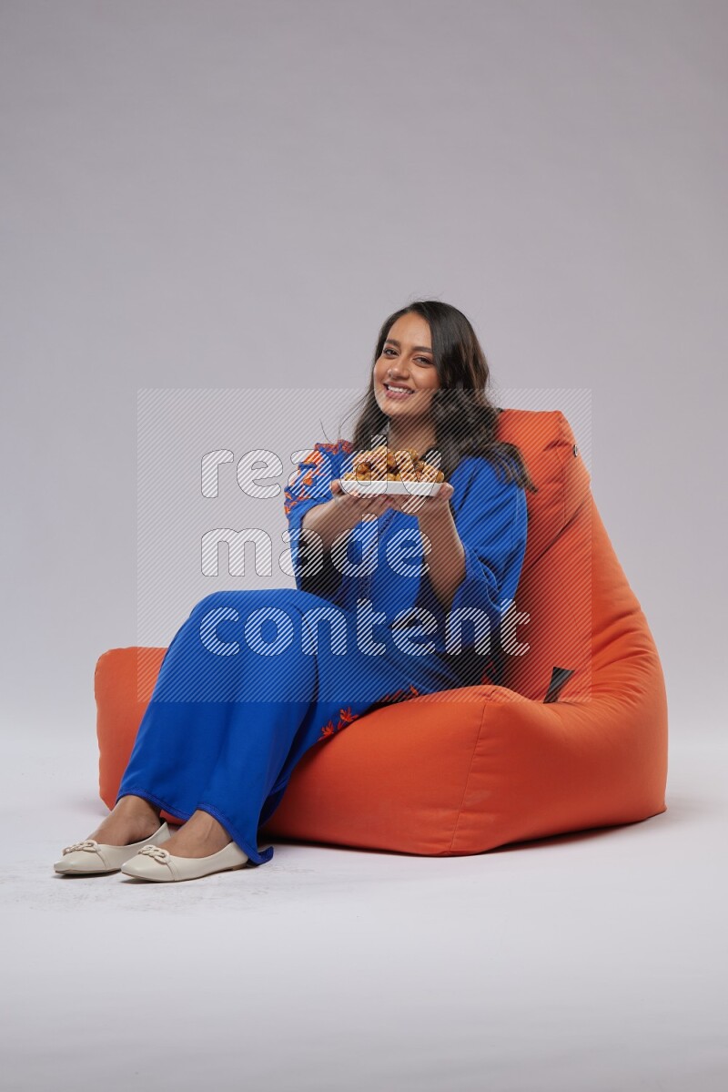 A Woman sitting on an orange beanbag wearing Jalabeya holding a plate of dates