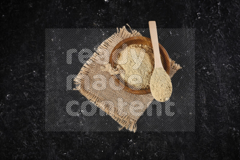 A wooden bowl full of ground ginger powder with a wooden spoon on it all on a burlap fabric on black background