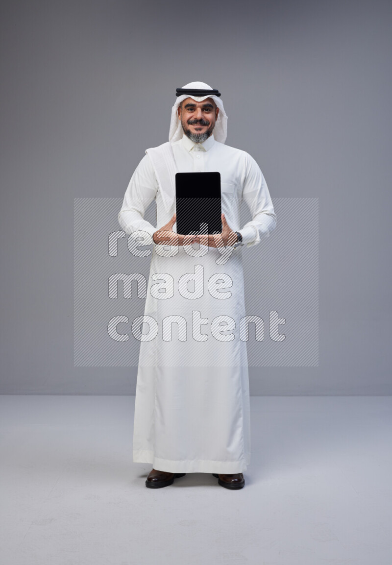 Saudi man Wearing Thob and white Shomag standing showing tablet to camera on Gray background