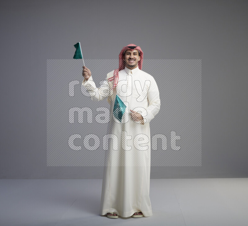 A Saudi man standing wearing thob and red shomag raising small Saudi flag on gray background
