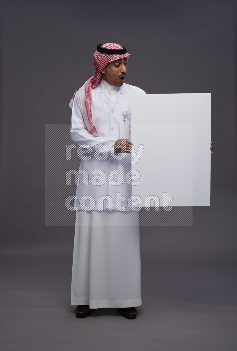 Saudi man wearing thob with lab coat and shomag with pocket employee badge standing holding board on gray background