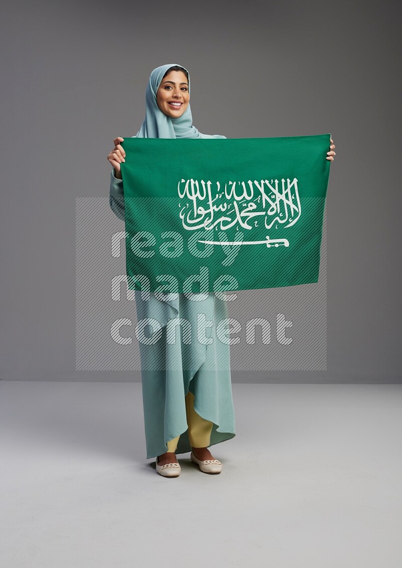 Saudi Woman wearing Abaya standing holding Saudi flag on Gray background