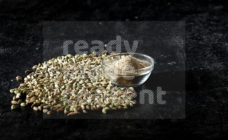 A glass bowl full of cardamom powder and cardamom seeds spreaded beneath the bowl on textured black flooring