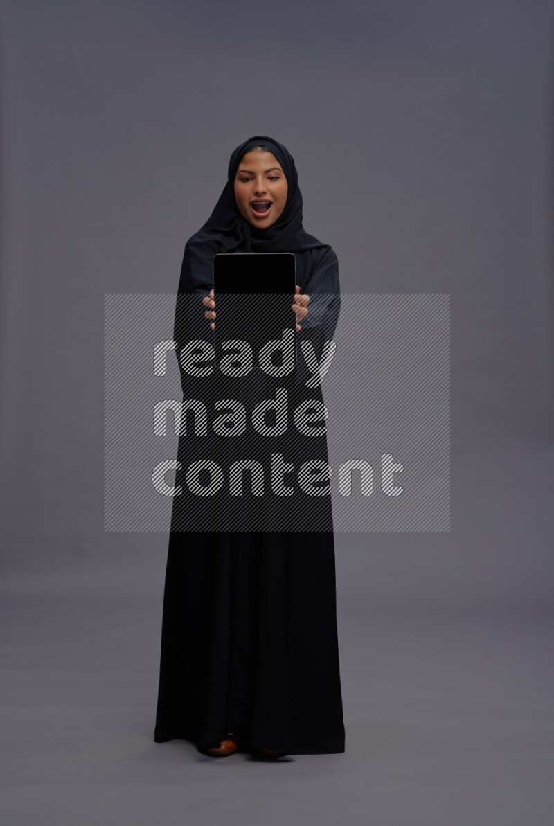 Saudi woman wearing Abaya standing showing tablet to camera on gray background