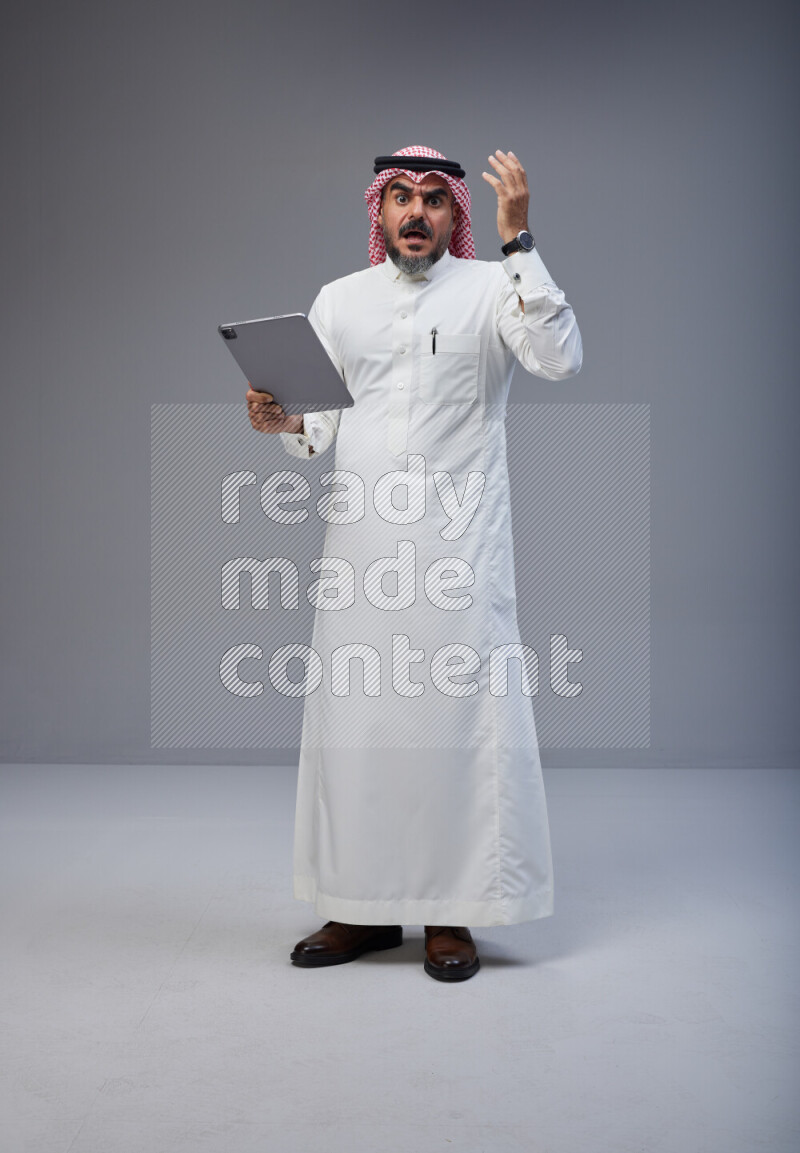 Saudi man Wearing Thob and red Shomag standing working on tablet on Gray background