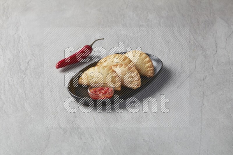Three fried sambosas in an oval shaped black plate and a sauce in a black round ramekin on a gray background