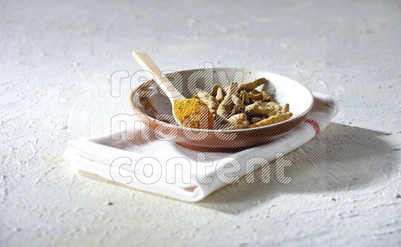 A plate filled with dried turmeric whole fingers and a wooden spoon full of turmeric powder on a textured white flooring