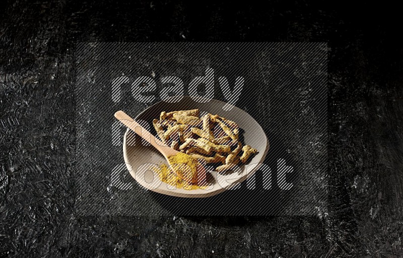 A plate filled with dried turmeric fingers and a wooden spoon full of turmeric powder on a textured black flooring