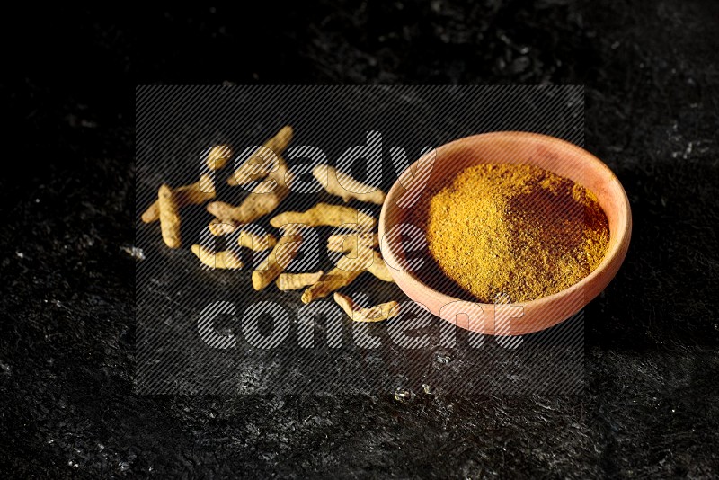 A wooden bowl full of turmeric powder with dried turmeric fingers on textured black flooring