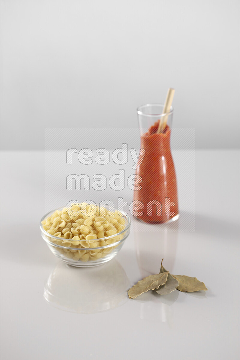 Raw pasta with tomatoe pasta with different ingredients such as cherry tomatoes, basil, garlic, bay laurel, cardamom, white pepper, black pepper, red chilis and wheat stalks on light grey background