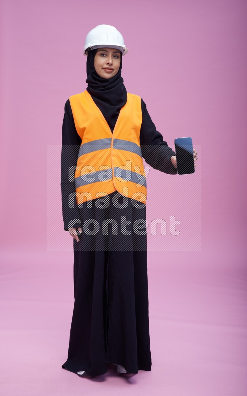 Saudi woman wearing Abaya with engineer vest and helmet standing showing phone to camera on pink background