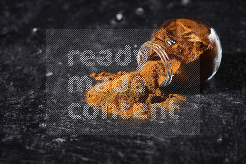 A glass jar full of ground paprika powder flipped with some spilling powder on black background