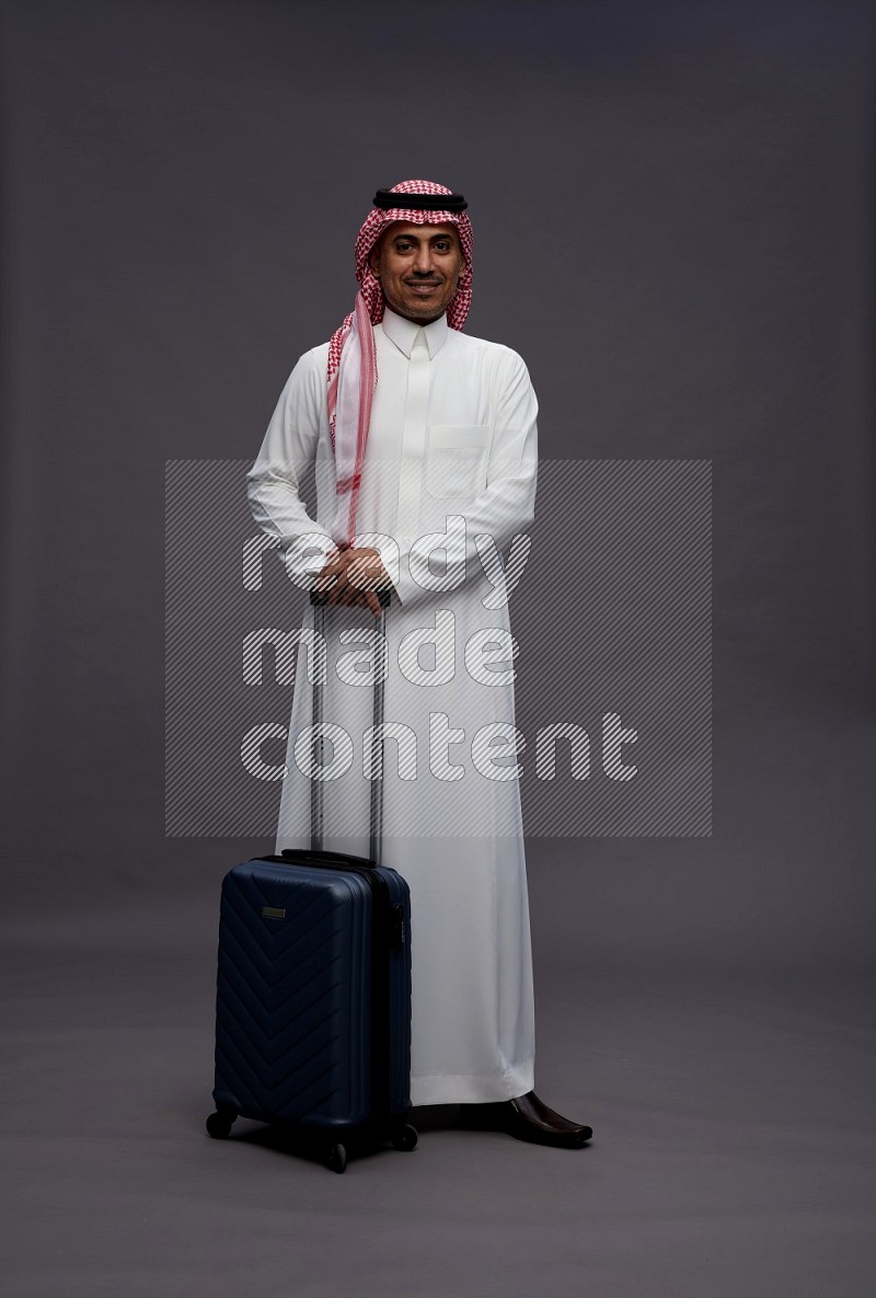 Saudi man wearing thob and shomag standing holding bag on gray background