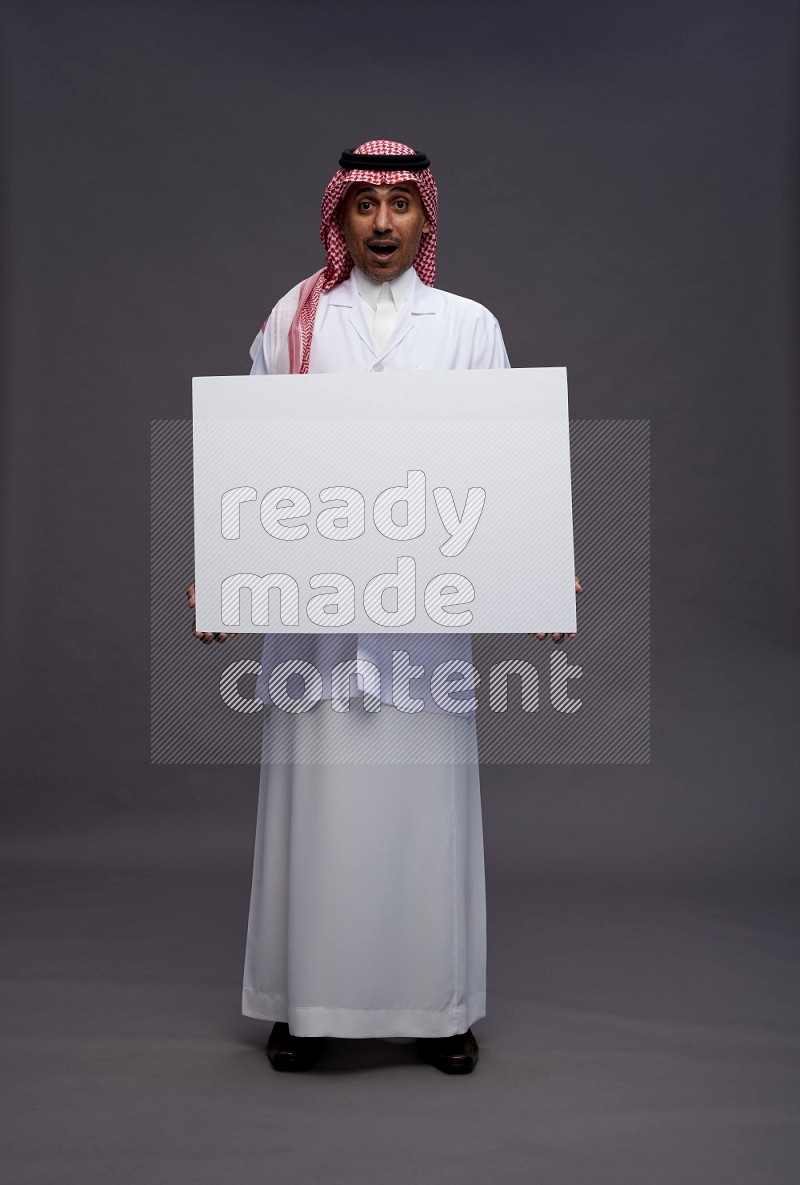 Saudi man wearing thob with lab coat and shomag with pocket employee badge standing holding board on gray background