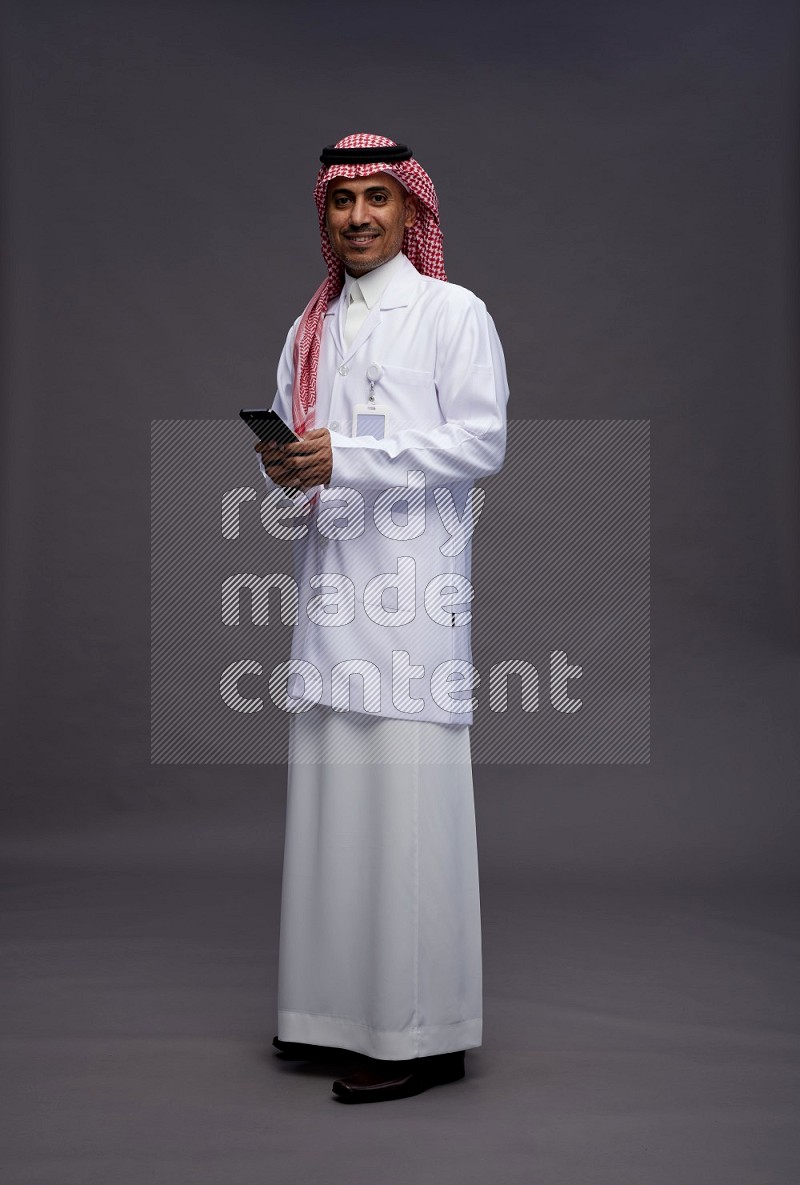 Saudi man wearing thob with lab coat and shomag with pocket employee badge standing texting on phone on gray background