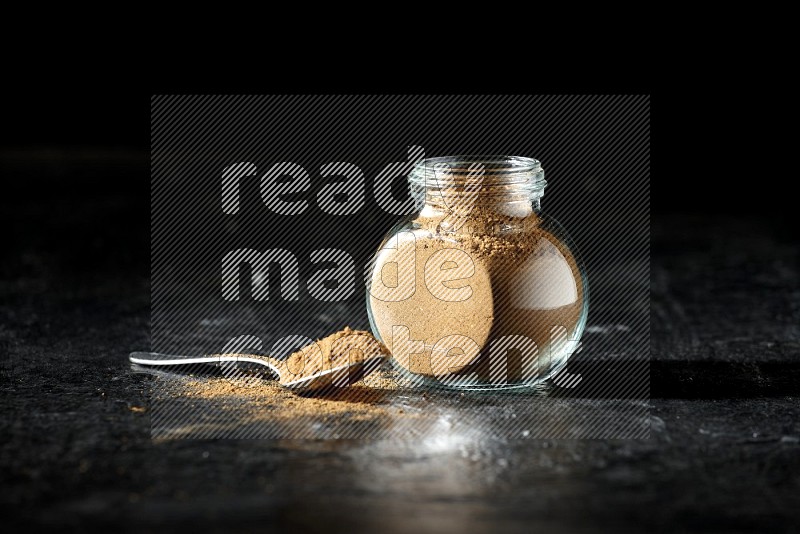 A glass spice jar and metal spoon full of allspice powder on a textured black flooring