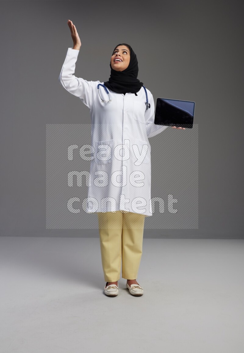 Saudi woman wearing lab coat with stethoscope standing showing tablet to camera with sign in the back on Gray background
