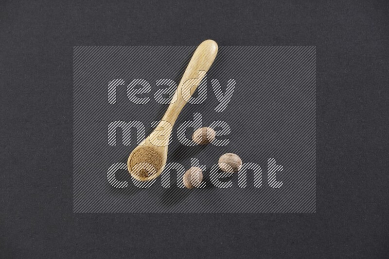 A wooden spoon full of nutmeg powder with whole nutmeg seeds beside it on a black flooring