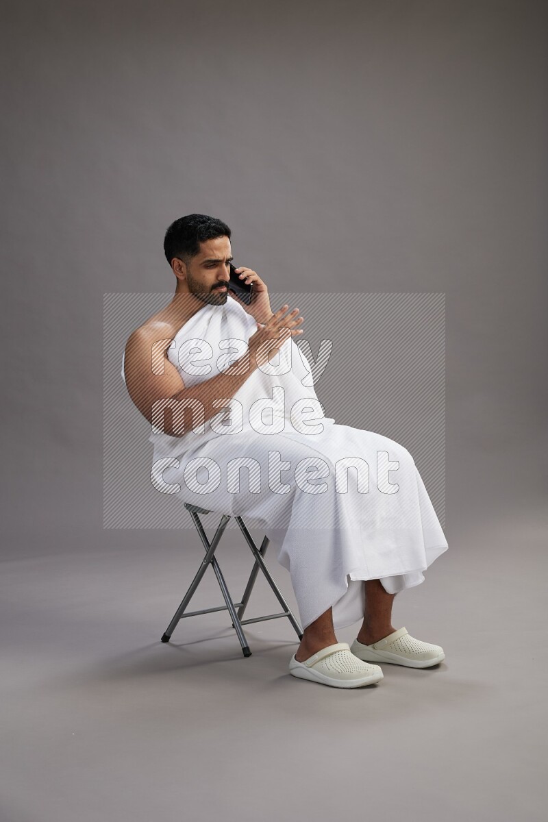 A man wearing Ehram sitting on chair talking on the phone on gray background
