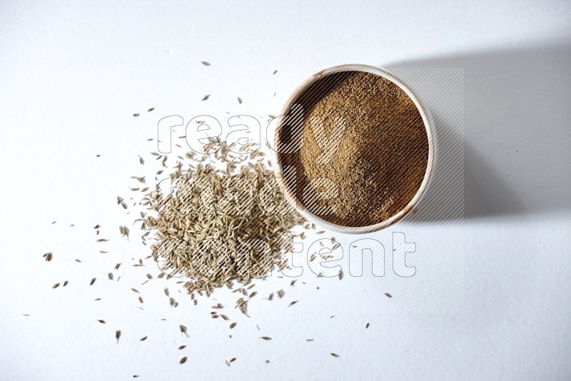 A beige bowl full of cumin powder with cumin seeds beneath it the bowl on a white flooring