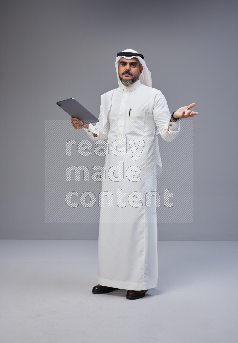 Saudi man Wearing Thob and white Shomag standing working on tablet on Gray background