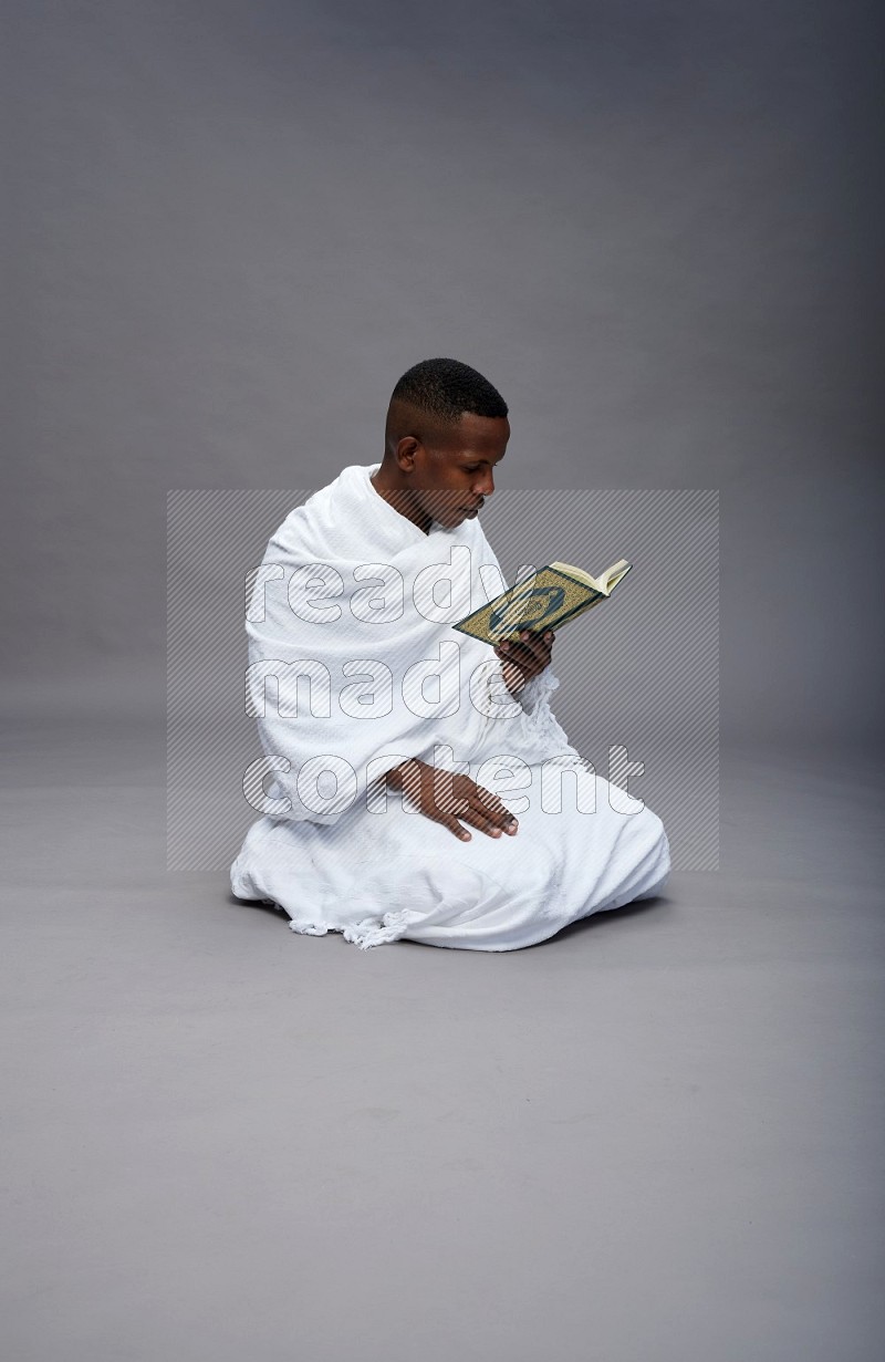 A man wearing Ehram sitting on floor reading quran on gray background