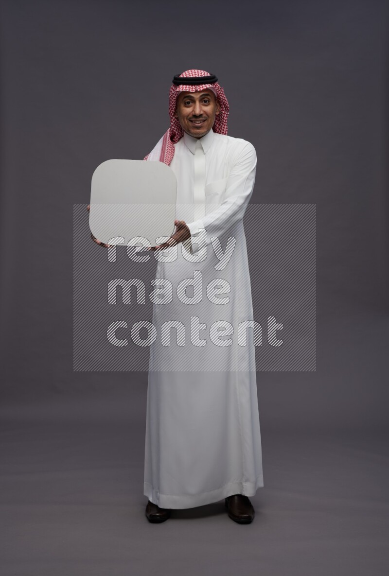 Saudi man wearing thob and shomag standing holding social media sign on gray background
