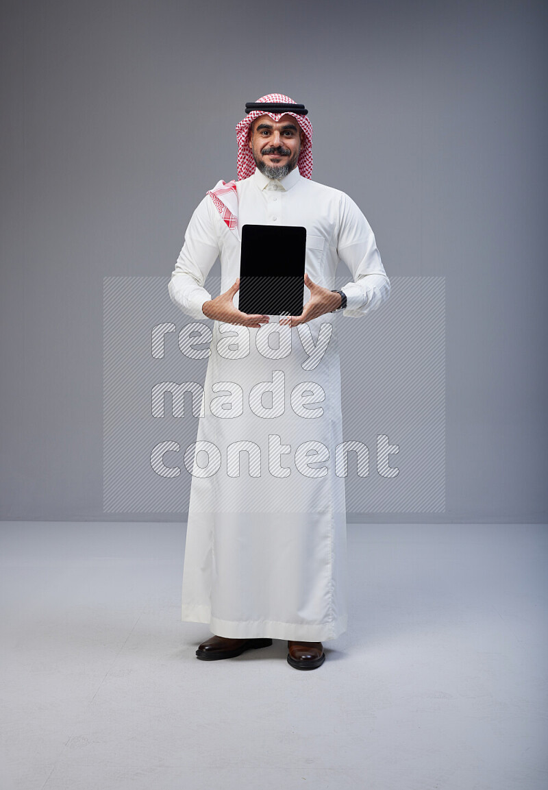 Saudi man Wearing Thob and red Shomag standing showing tablet to camera on Gray background