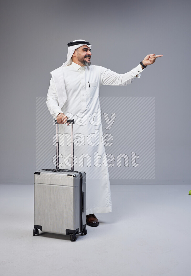 Saudi man wearing Thob and white Shomag standing holding Travel bag on Gray background