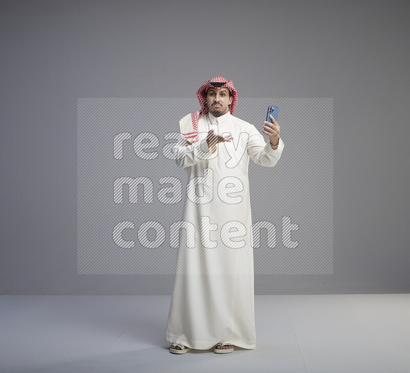A Saudi man standing wearing thob and red shomag taking selfie on gray background