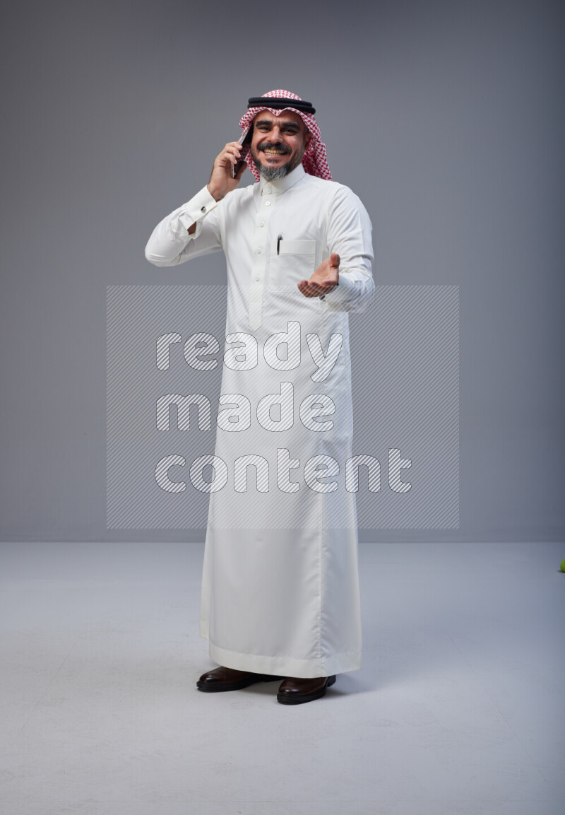 Saudi man Wearing Thob and red Shomag standing talking on phone on Gray background