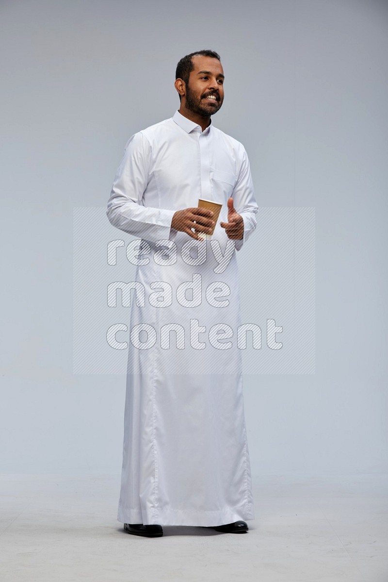 Saudi man wearing thob standing holding paper cup on gray background