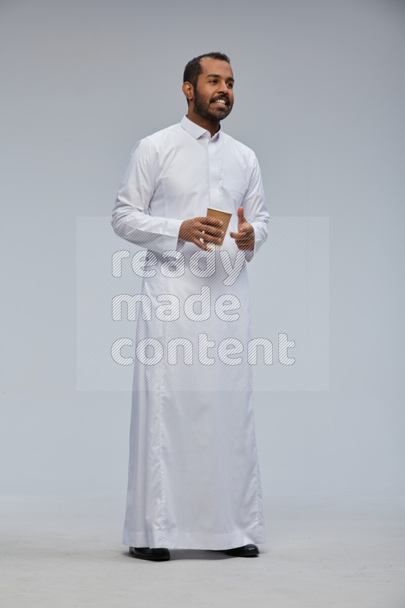 Saudi man wearing thob standing holding paper cup on gray background