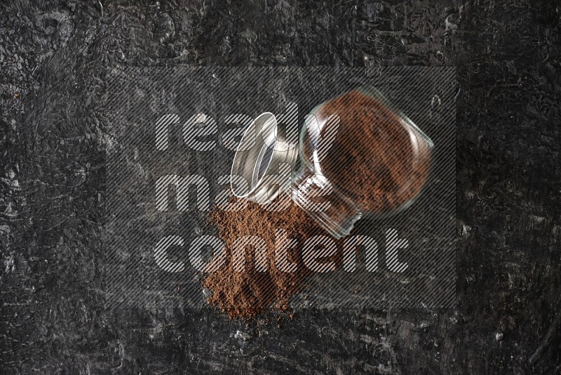 A flipped glass spice jar full of cloves powder and powder came out of it on textured black flooring